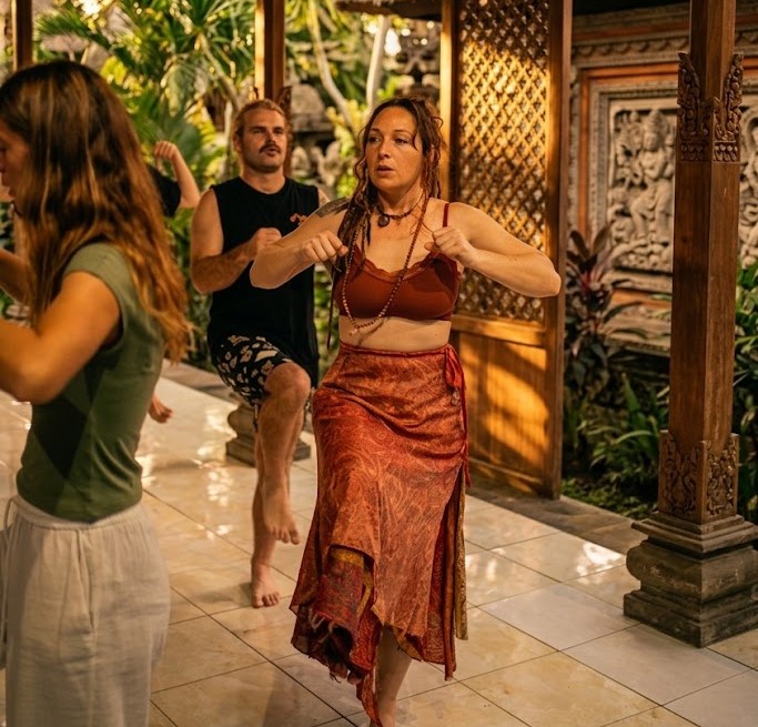 A somatic breathwork session in Bali at golden hour; participants are in active, standing release poses with focused expressions, set against a backdrop of traditional stone carvings and tropical palms.